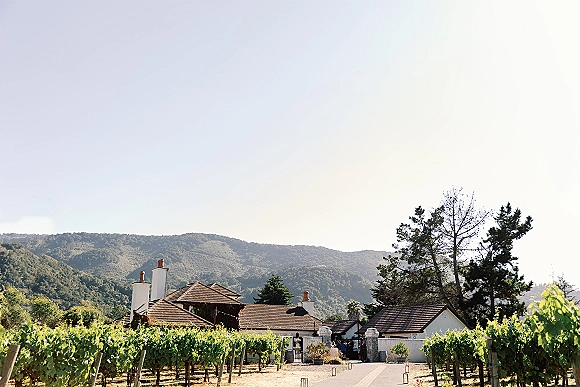 Vineyard wedding venue entrance with winery buildings and a stone gate, walkway through grapevines toward mountain hills under open sky