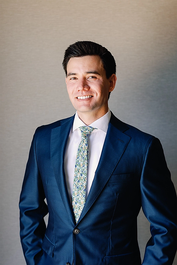 Groom portrait in a navy blue wedding suit with a floral tie, standing against a neutral wall in soft indoor light