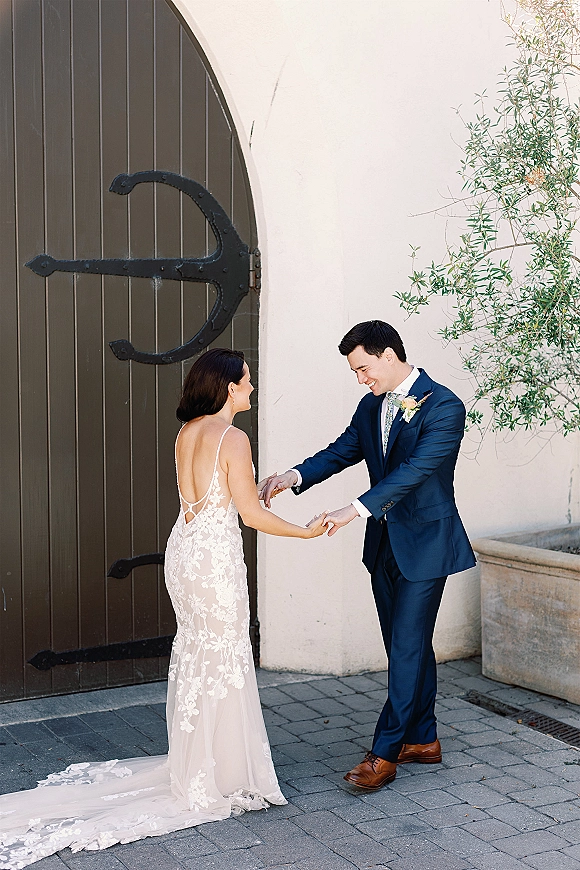 First look moment as bride in a lace wedding dress touches groom’s hand by an arched wooden door, his reaction candid against stucco wall