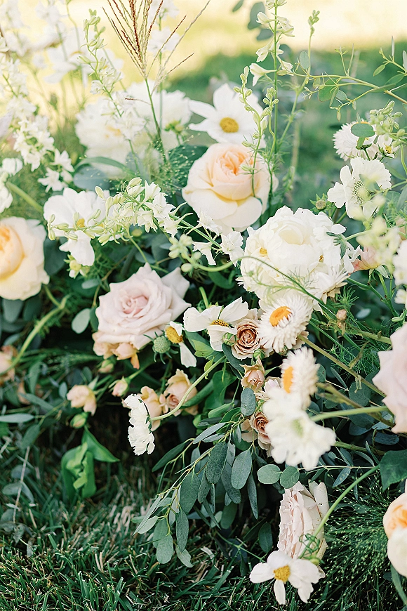 Wedding florals with white and blush roses, daisies, and eucalyptus arranged in a meadow-like display on a grass lawn in a garden setting