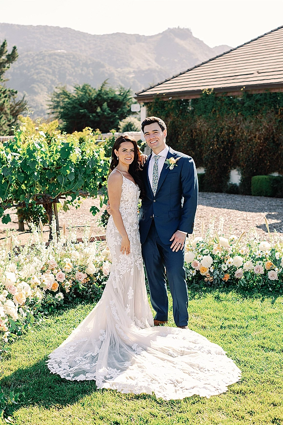 Couple portrait of bride in a lace gown with long train and groom in suit, arm around her in vineyard rows with mountains behind