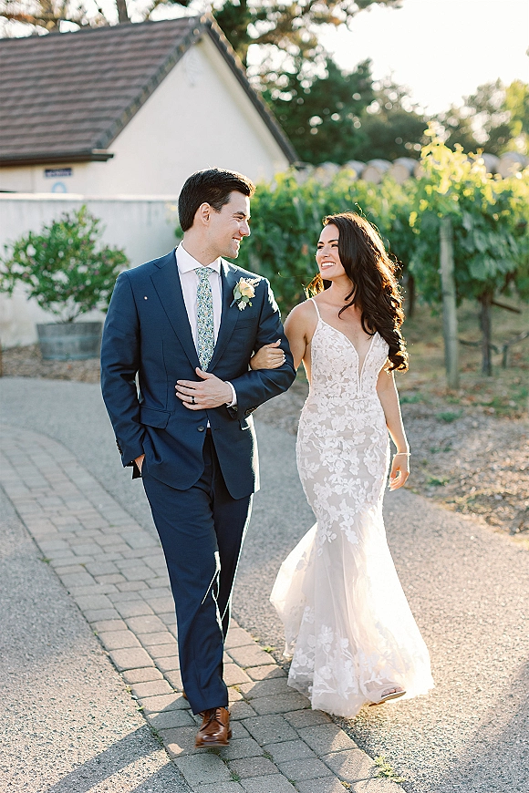 Couple portrait of bride and groom walking arm in arm, her lace wedding dress beside his blue suit along sunlit vineyard rows