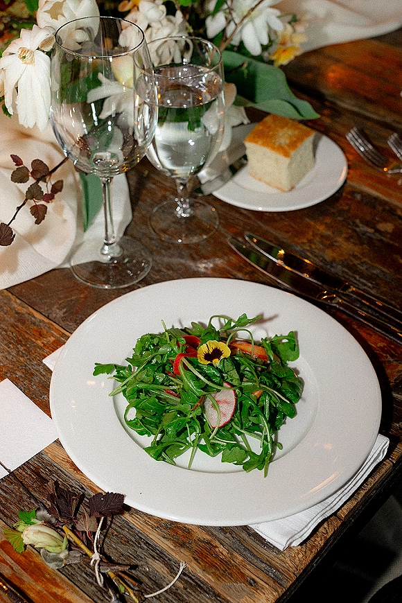 Reception tablescape with white dinner plates, salad course and wine glasses on a rustic wooden table, finished with a greenery centerpiece