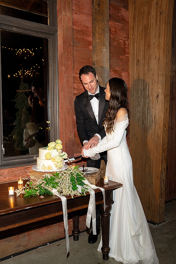 Wedding cake cutting as bride and groom slice a two-tier buttercream cake with floral topper on a candlelit rustic table indoors