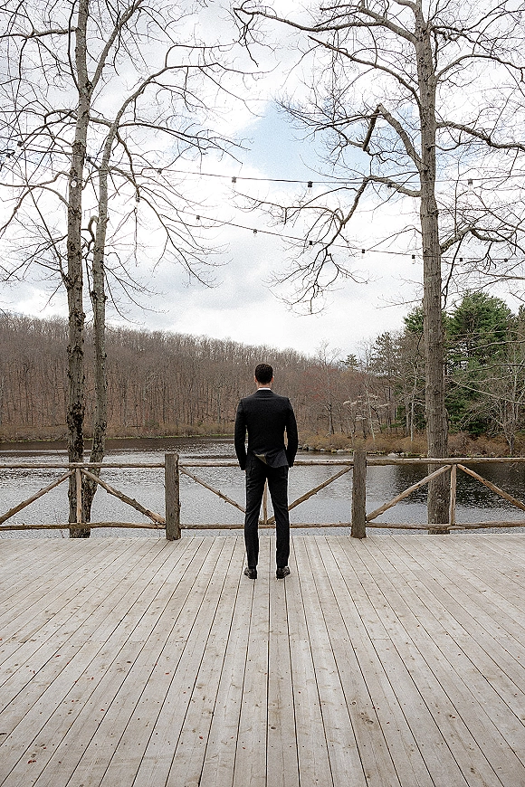 Groom portrait of a black suit groom from behind on a wooden deck, looking at a lake under string lights and cloudy sky