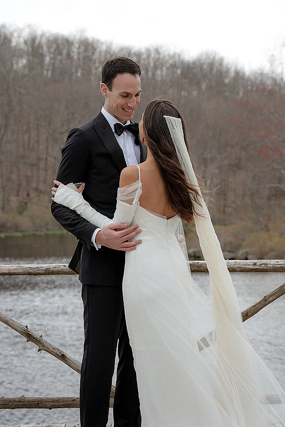 Couple portrait of bride and groom embrace by a lake, her veil blowing in the wind as they smile beside a wooden fence