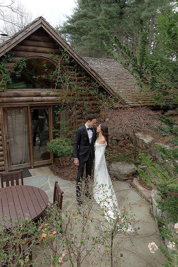 Couple portrait of bride in an off-the-shoulder wedding dress and groom in a tuxedo, forehead touching on a stone patio by a log cabin