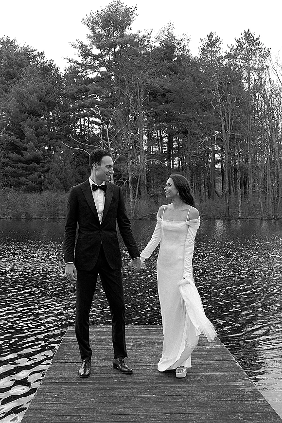 Couple portrait in a black and white wedding photo, bride in long sleeve dress and groom in tuxedo walking hand in hand on a lake dock