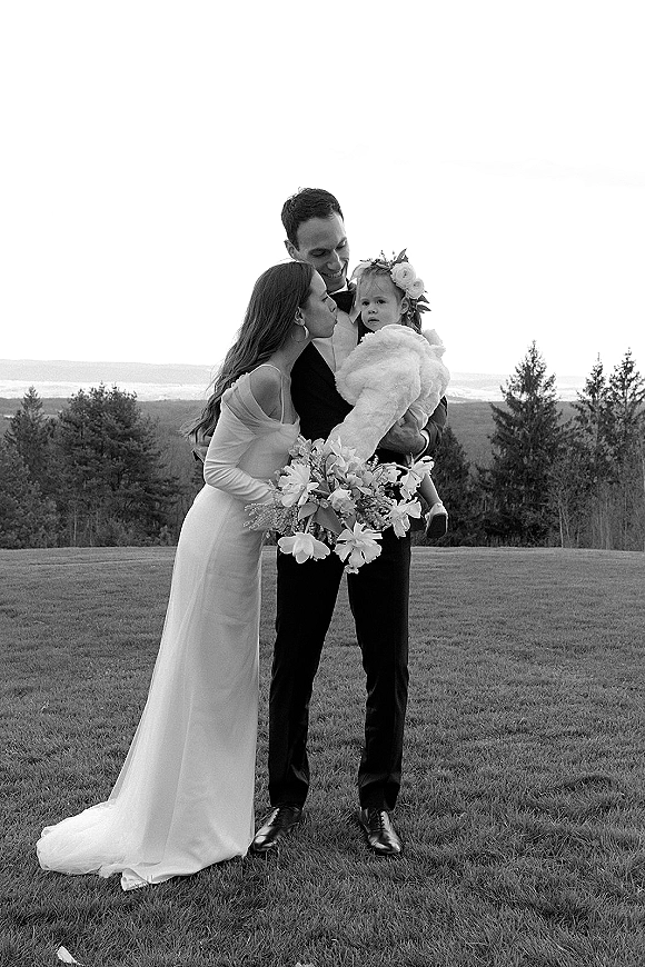Couple portrait of bride kissing groom as he holds their child, bride with bouquet, in a grassy field with trees and distant hills