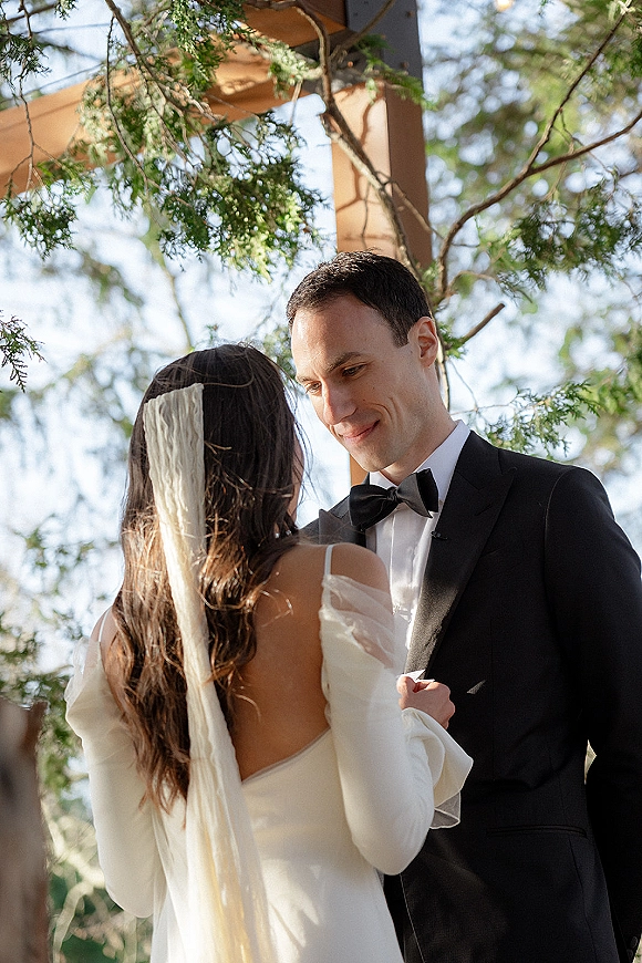 Wedding vows as bride in off-the-shoulder dress and long veil faces groom in bow tie under a wooden arbor with trees and sky behind