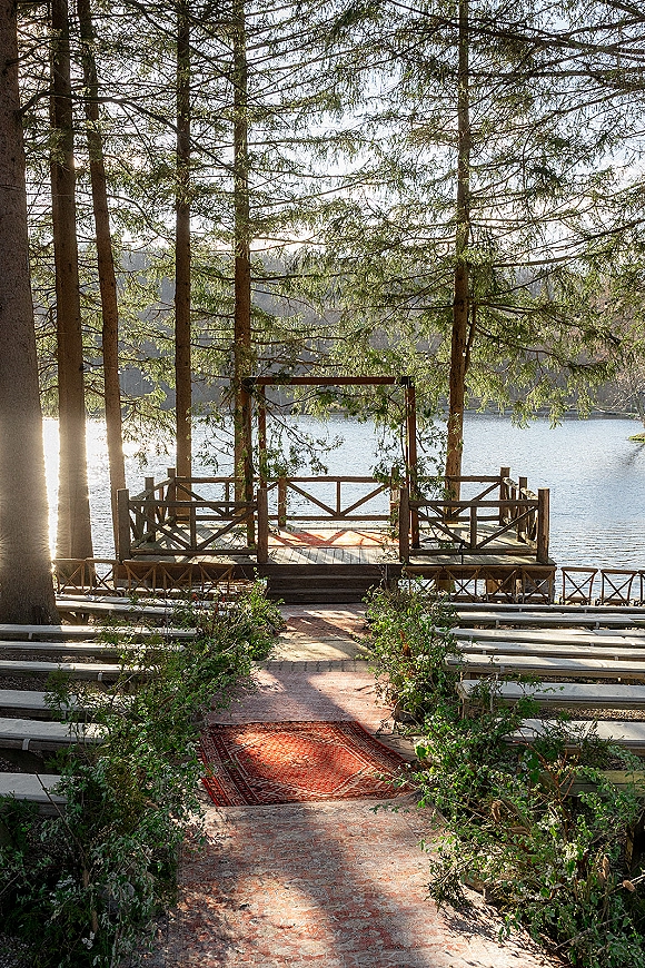 Ceremony setup for an outdoor wedding ceremony with a wood arch, rug-lined aisle and greenery, on a deck overlooking a sunlit lake and forest
