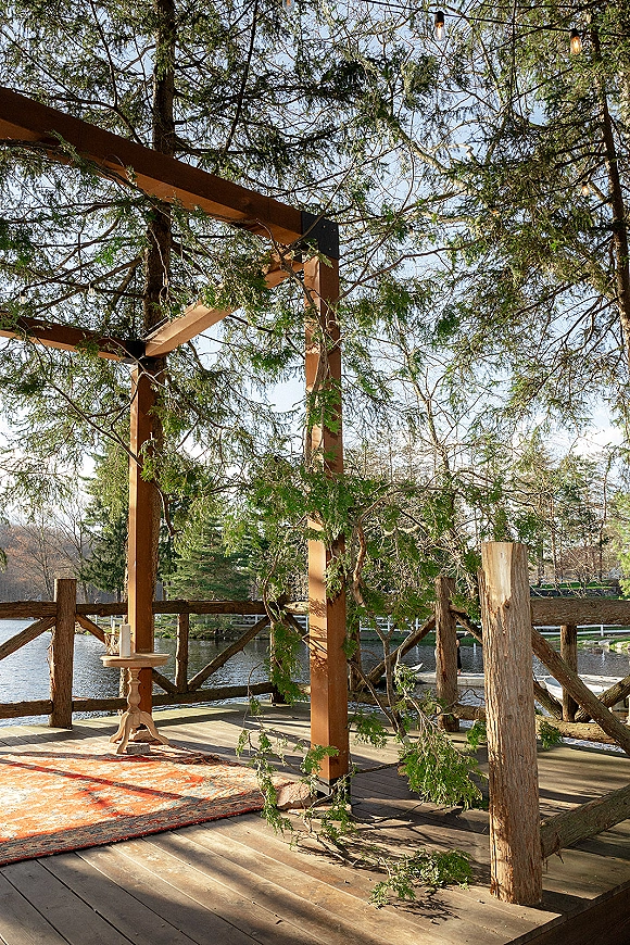 Ceremony altar with wood pergola altar, evergreen garland and string lights on a deck overlooking a lake, with rug and candles