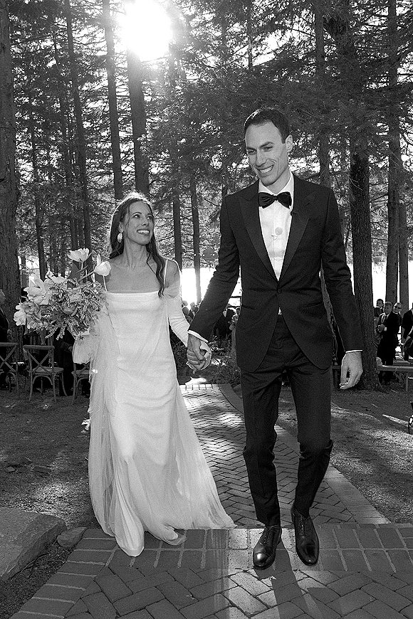 Wedding recessional as bride and groom walking hand in hand down a brick path, bouquet and veil catching sunlight with forest trees behind