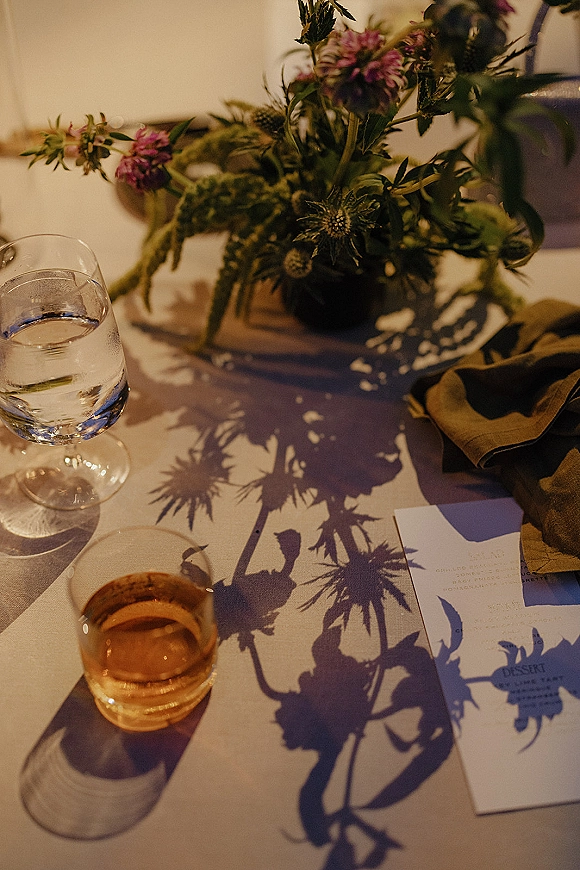Wedding tablescape with reception table centerpiece of wildflowers and greenery, bud vase, menu and place cards, warm lighting on white linens
