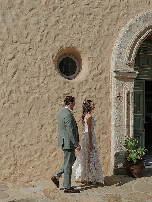 Couple portrait of bride in a lace gown and groom in a green suit walking past a stone arch doorway with stucco wall backdrop