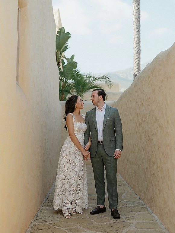 Couple portrait of bride and groom walking hand in hand, bride in a lace wedding dress beside his green suit, with stucco walls and palms