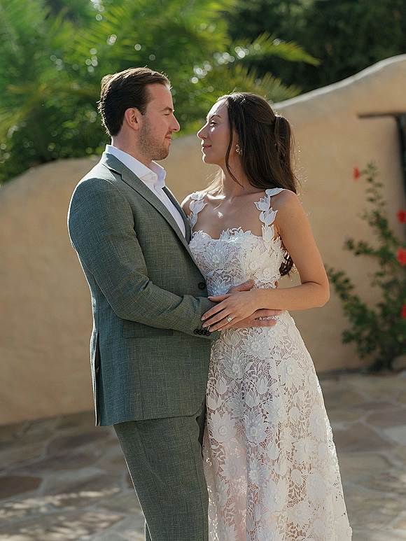 Couple portrait of bride and groom embrace, gazing at each other; bride in lace dress beside stucco wall with palm trees behind