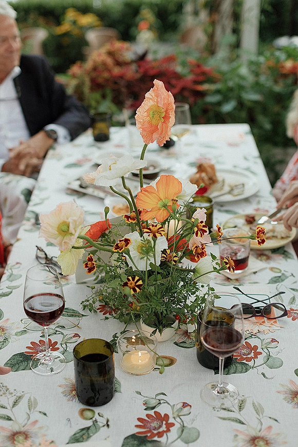 Reception tablescape with a wildflower wedding centerpiece of poppies and marigolds, red wine, green tumblers and votive candles in a garden setting