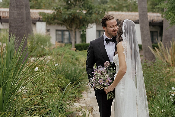 Couple portrait of bride and groom in close embrace, bride holding a bouquet with cathedral veil, on a garden path by palms and stucco building