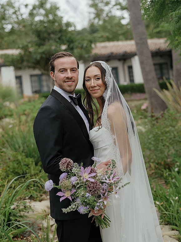 Couple portrait of bride and groom holding a lavender bouquet, bride in strapless dress with lace veil in a garden setting
