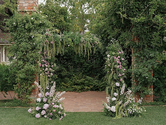 Wedding ceremony arch with hanging greenery and pink, white, and purple flowers on brick pillars, framed by lush garden lawn backdrop