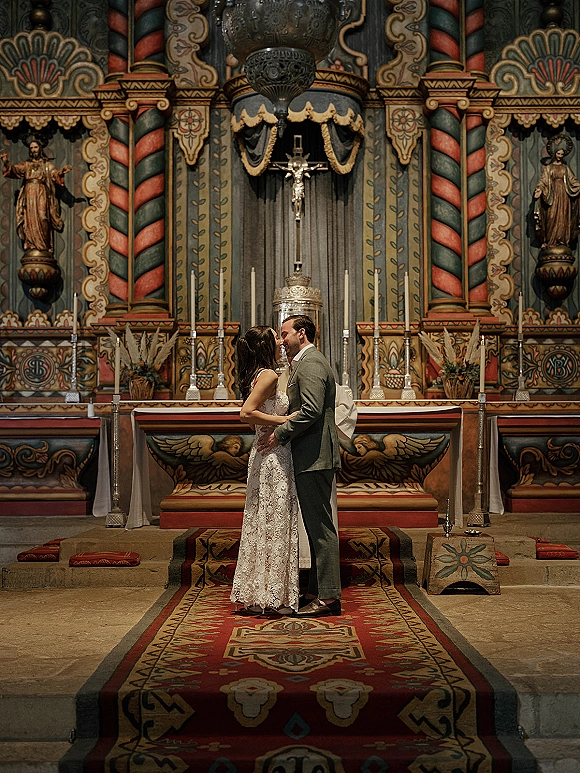 Wedding kiss at the altar as bride in a lace dress and veil embraces groom, framed by crucifix, candles, and ornate church interior