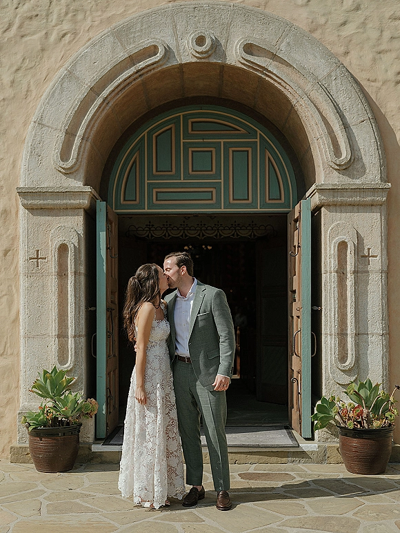 Wedding kiss portrait of bride and groom kissing in a stone arch church doorway, bride in lace dress with succulent accents nearby