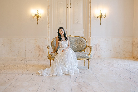 Bridal portrait of a bride sitting on couch in a strapless beaded lace gown with long train on marble floor beside wall sconces