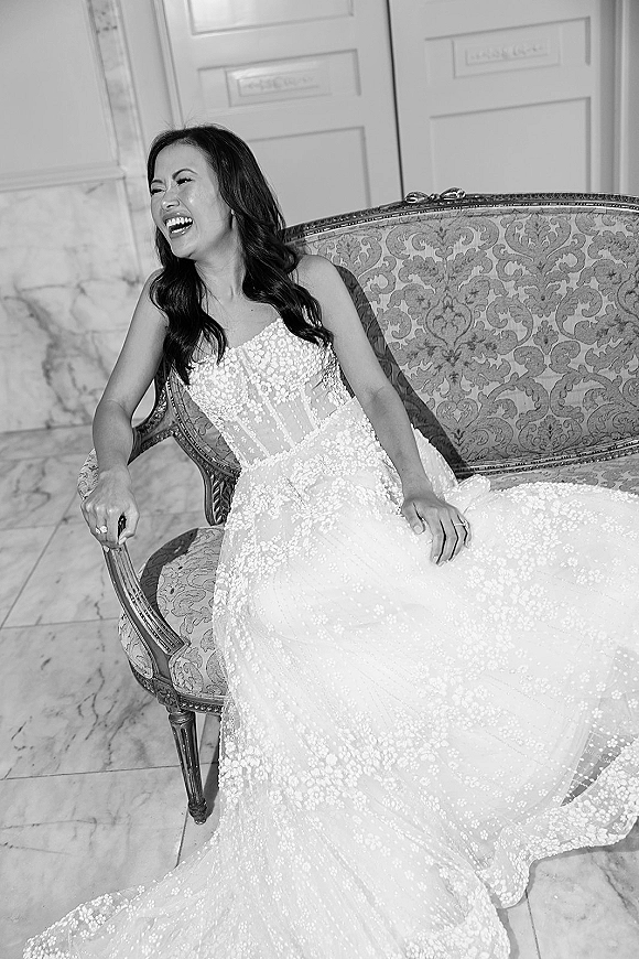 Bridal portrait of a laughing bride in a strapless beaded lace gown on an upholstered settee, showing her wedding ring on a marble floor