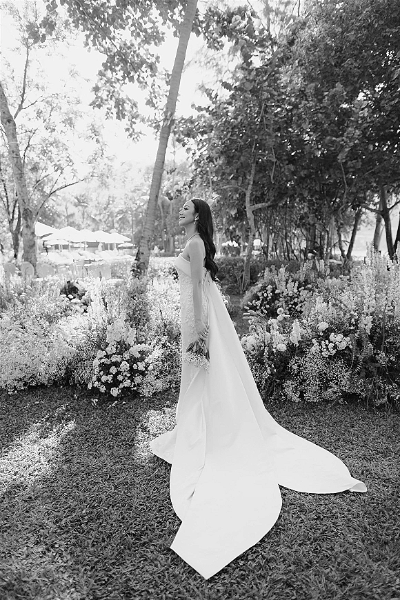 Bridal portrait in black and white of a bride in side profile holding a bouquet, wearing a strapless dress with long train in a garden setting