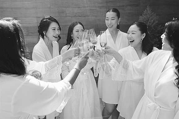 Bridesmaids toast with champagne flutes during a getting ready toast, wearing lace-trim robes and earrings against a wood wall with greenery