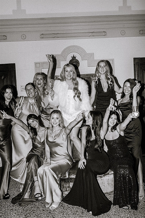 Bridal party portrait of the bride with bridesmaids raising champagne flutes in satin and sequin gowns by a fireplace mantel indoors