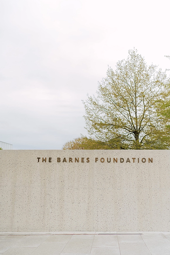 Venue exterior with wedding venue entrance concrete wall signage in metal lettering, set by stone paving with trees and sky behind