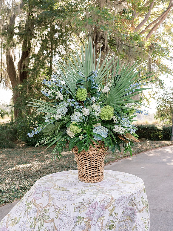 Wedding floral arrangement with palm fronds, hydrangea, and blue flowers in a woven basket on a table along a mossy garden path