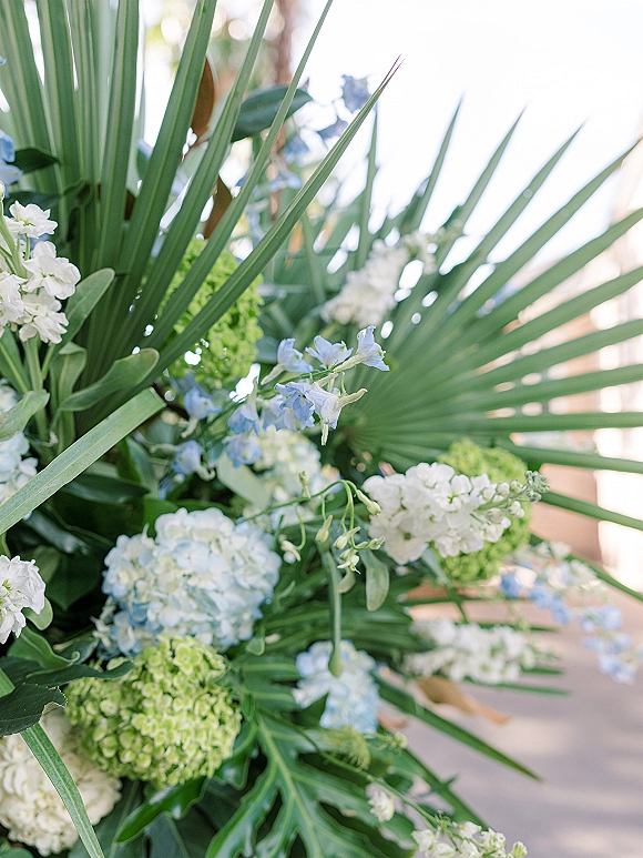 Wedding floral arrangement with tropical wedding flowers and palm fronds, blue and white blooms on wood decking in bright outdoor light
