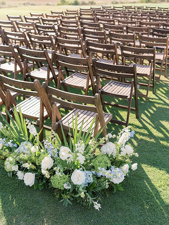 Ceremony seating with wood folding chairs in neat rows, lined by low white rose and hydrangea aisle florals on a grass lawn
