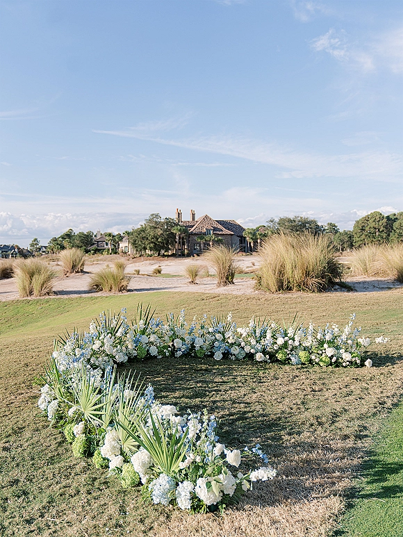 Ceremony aisle florals with grounded aisle flowers in white and blue hydrangea, palm fronds, and greenery along a lawn by sand dunes under blue sky