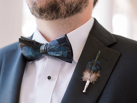 Groom accessories featuring a blue feather bow tie and feather boutonniere on a black tuxedo with white shirt against a soft neutral background