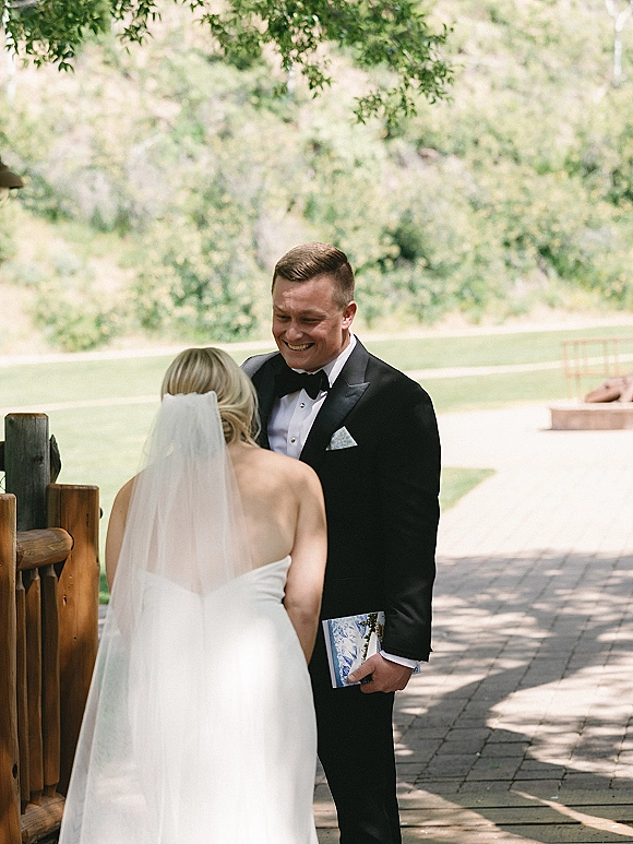 First look moment as bride in veil and strapless dress approaches groom in black tux holding a program on a garden walkway by wooden fence