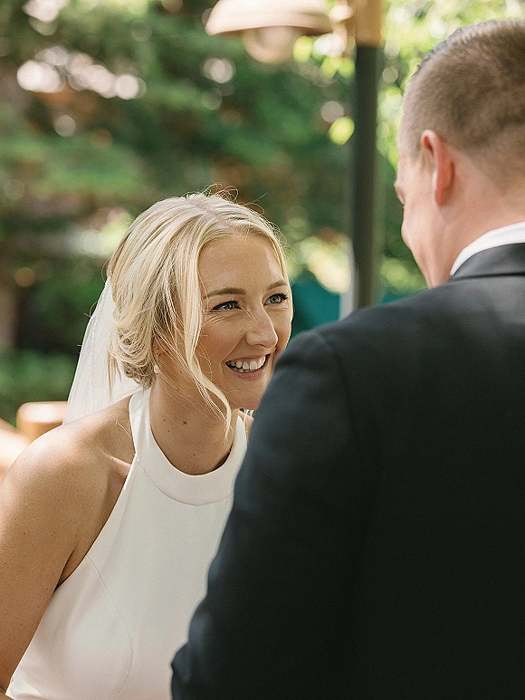 Ceremony moment as bride smiling at groom in a halter wedding dress and veil, with bistro lights and garden trees behind them