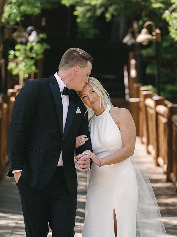 Couple portrait of bride and groom kiss as he kisses her forehead on a wooden bridge, her veil draped over his tuxedo amid trees