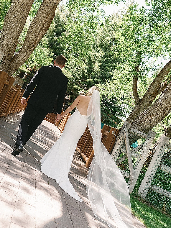 Bride and groom walking away hand in hand, her long veil trailing behind, along a tree-lined brick path with dappled sunlight