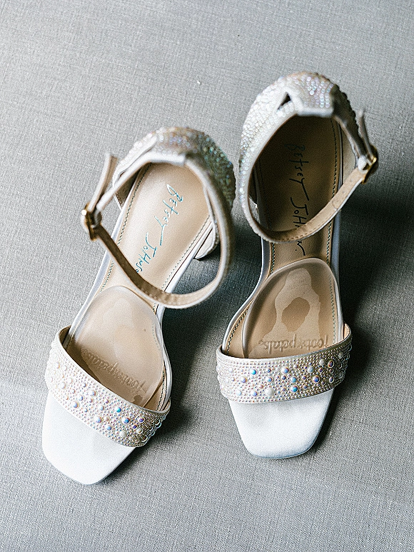 Bridal shoes with white bridal heels featuring rhinestone beading and satin ankle straps on a soft gray fabric surface flat lay