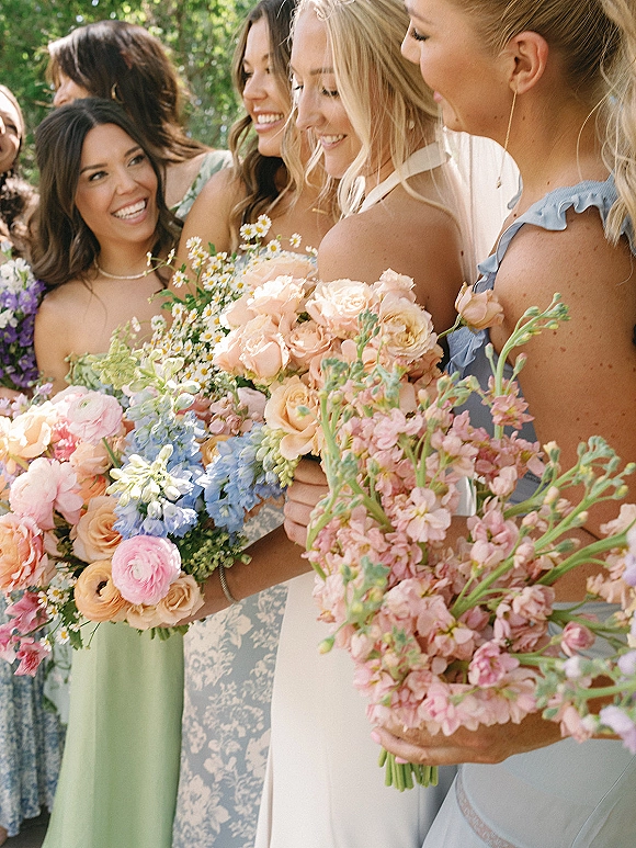 Bridesmaid group photo with bridesmaids holding bouquets of blush roses, blue delphinium and daisies in sunlit greenery outdoors