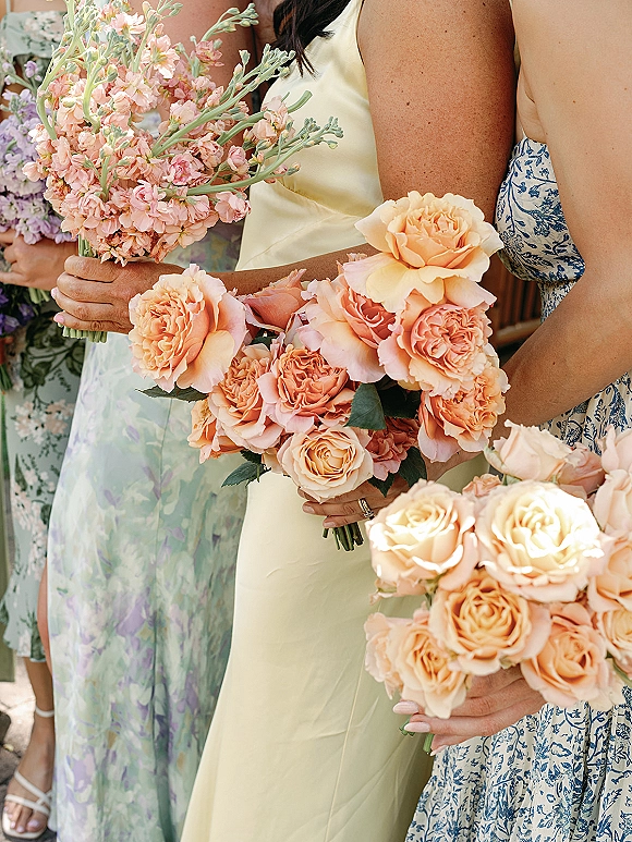 Bridesmaid bouquets of peach rose bouquet blooms held by women in pastel dresses, one showing a wedding ring, on a sunlit stone walkway
