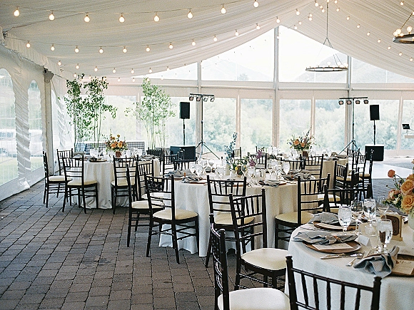Reception tablescape with round wedding reception tables, white linens, black Chiavari chairs, floral centerpieces under a clear-top tent with string lights