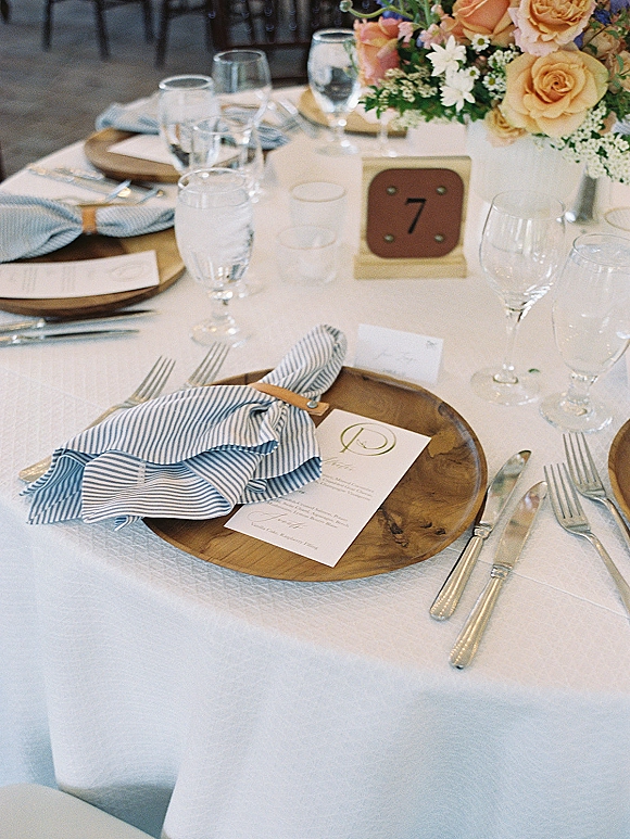 Reception tablescape with wedding place setting on wood charger plates, striped napkins, menu cards, and rose-daisy centerpiece in reception room