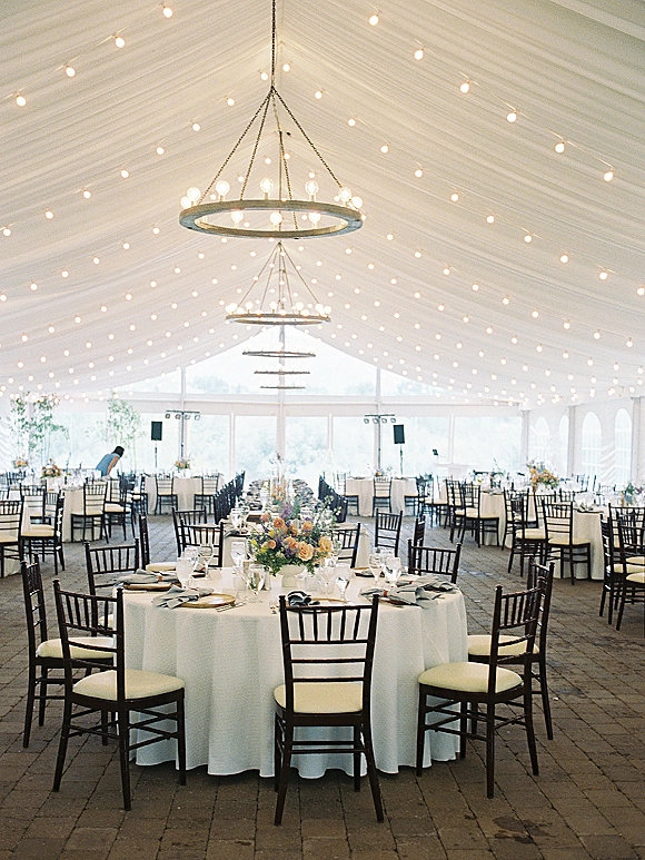 Wedding reception decor with string lights and round chandeliers over white tablecloth round tables in a clear-walled tent on brick pavers
