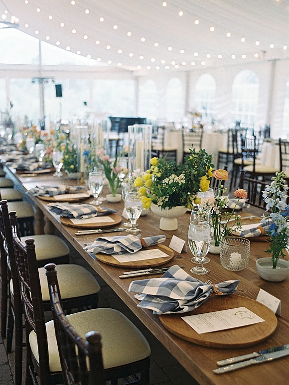Reception tablescape on a long farm table wedding setup with crossback chairs, greenery, wildflower centerpieces, and candles under a white tent
