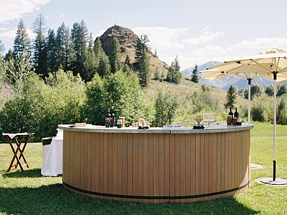 Outdoor wedding bar with a round wooden counter, wine bottles, glassware and dispensers under patio umbrellas, set on a mountain lawn